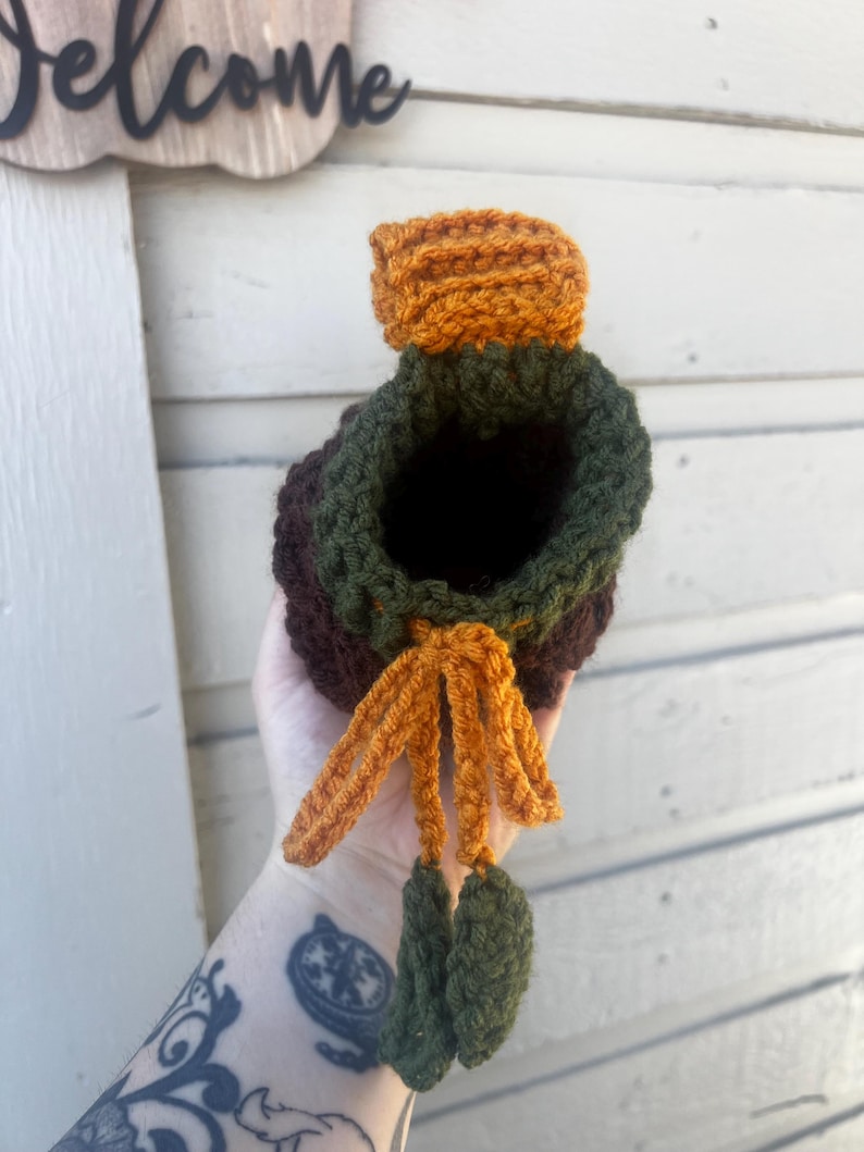 May include: A crochet drawstring pouch featuring a dark brown base, a forest green upper section, and a bright orange top flap. The pouch includes an orange drawstring cord finished with two small, dark green crocheted leaf-shaped accents.