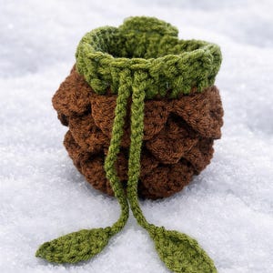 May include: A small, round, crocheted drawstring pouch featuring a textured brown body resembling a pinecone and a green top rim. The pouch includes two long green crocheted drawstrings that hang down, each finished with a small green leaf-shaped tip.