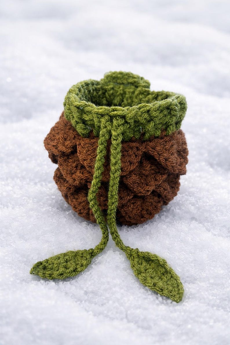 May include: A small, round, crocheted drawstring pouch featuring a textured brown body resembling a pinecone and a green top rim. The pouch includes two long green crocheted drawstrings that hang down, each finished with a small green leaf-shaped tip.