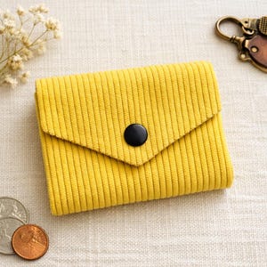 May include: A small, yellow corduroy wallet with a black button closure. The wallet has a flap closure and a vertical ribbed texture. A few coins and a keychain are visible in the background.