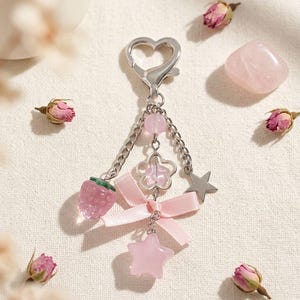 Könnte beinhalten: Ein silberfarbener Schlüsselanhänger mit herzförmigem Verschluss, verziert mit einem rosa Erdbeer-Anhänger, einem Blumen-Anhänger, einem Stern-Anhänger und einer rosa Schleife. Der Schlüsselanhänger hat auch rosa Perlen und einen kleinen sternförmigen Anhänger. Getrocknete Rosenknospen und ein rosa Kristall sind verstreut.