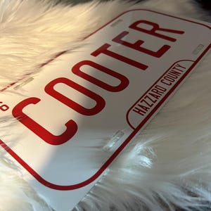May include: White license plate with the word "COOTER" in bold red letters. The text "HAZZARD COUNTY" is printed in red below. The plate has a red border and is resting on a white, fluffy surface.
