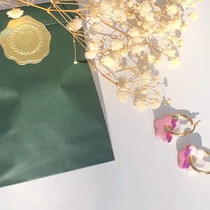 Op de afbeelding: Een paar gouden oorringen met roze en witte bloemvormige bedels. De oorbellen liggen op een wit oppervlak naast een donkergroene verpakking, verzegeld met een gouden sticker en droogbloemen.