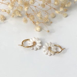 Könnte beinhalten: Ein Paar goldene Creolen mit weißen Gänseblümchen-Anhängern. Die Ohrringe sind vor einem weißen Hintergrund mit getrockneten Blumen im oberen Teil des Bildes platziert. Ein zartes und feminines Accessoire.