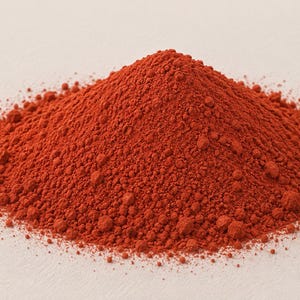May include: A conical mound of fine, vibrant red powder with a granular texture. The substance is composed of tiny, uniform particles that form a dense, rounded peak, displaying a consistent deep crimson colour throughout the entire pile.