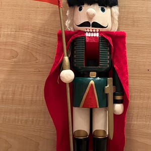 May include: A wooden nutcracker figurine wearing a tall black fuzzy hat, a red velvet cape, and a green jacket with gold accents. The figure holds a tall gold pole with a red triangular flag and has a small sword attached to its side.