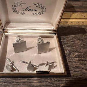 May include: A set of silver-toned square cufflinks and matching shirt studs housed in a cream-colored Anson presentation box. A wooden ruler is visible in the background, showing measurements in inches along the top edge of the box.
