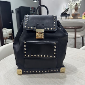 Puede incluir: Mochila negra con tachuelas y detalles dorados. La solapa presenta un logotipo y se asegura con un cierre dorado. La bolsa tiene un asa superior y un bolsillo frontal.