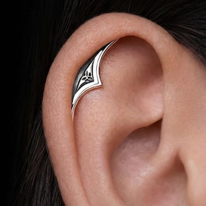 Peut inclure: Un ear cuff argenté avec un motif géométrique, orné d'un motif floral noir et blanc. Le cuff est incurvé pour s'adapter au cartilage de l'oreille. Le bijou est présenté sur l'oreille d'une personne, avec des cheveux foncés en arrière-plan.