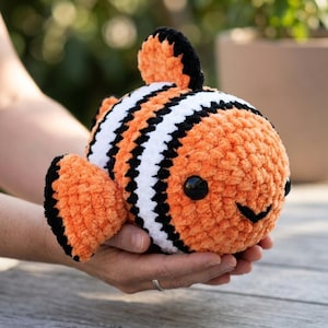 Könnte beinhalten: Ein handgefertigtes Clownfisch-Spielzeug. Der runde, orangefarbene Körper hat schwarze und weiße Streifen, schwarze Flossen und schwarze Knopfaugen. Das weiche Spielzeug wird in offenen Händen gehalten.