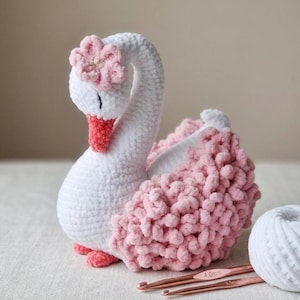 Puede incluir: Un cisne de ganchillo hecho a mano en blanco y rosa. El cisne presenta un cuerpo, cuello y cabeza blancos, con una flor rosa en la cabeza y detalles rosas en el pico y las patas. La espalda del cisne está adornada con hilo rosa esponjoso. Se ven ganchillos y ovillos de lana.