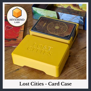 May include: A bright yellow rectangular plastic card case for the game Lost Cities. The lid features the embossed text LOST CITIES. The base holds a deck of cards with a dark brown back design featuring a gold circular emblem and thin lines.