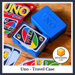 May include: A blue plastic travel case for a card game, featuring the embossed text LAURA'S UNO on the lid. The case holds a deck of cards with a visible WILD card. An original UNO game box and loose playing cards are visible nearby.
