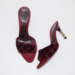 Pode incluir: Um par de sandálias deslizantes de veludo bordô. As sandálias apresentam bico arredondado e uma tira na parte superior com o padrão do logotipo Gucci. Os saltos têm um design de bambu dourado. Os sapatos estão sobre um fundo branco.