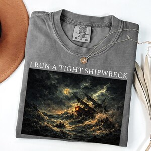 Könnte beinhalten: Graues T-Shirt mit dem Schriftzug "I RUN A TIGHT SHIPWRECK" über einer Grafik eines Schiffes in einem Sturm. Das Shirt hat einen Rundhalsausschnitt und kurze Ärmel. Eine silberne Halskette mit Anhänger befindet sich auf dem Shirt.