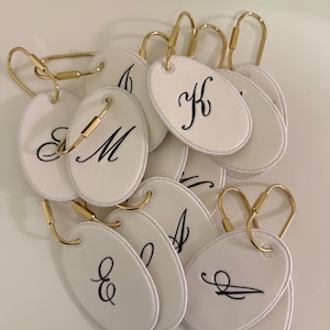Pode incluir: Uma coleção de etiquetas ovais de tecido branco, cada uma com uma letra preta bordada em letra cursiva. Cada etiqueta está presa a um fecho de argola de metal dourado. As letras visíveis incluem K, M, E, A e outras.