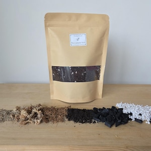 Könnte beinhalten: Ein beigefarbener Standbeutel mit der Aufschrift Verdant Leaf Terrarium Soil und einem Sichtfenster, das dunkle Erde zeigt. Davor liegen Häufchen aus Sand, getrocknetem Moos, Kokosfaser, dunkler Blumenerde, Holzkohle und weißen Perlit-Granulaten.