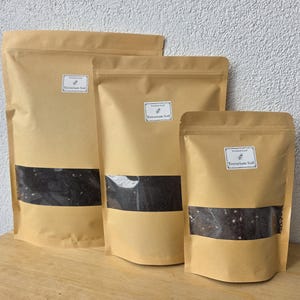 Könnte beinhalten: Drei Standbeutel in verschiedenen Größen aus hellbraunem Papier, jeder mit einem klaren rechteckigen Sichtfenster, das dunkle Erde mit weißen Partikeln zeigt. Jeder Beutel hat ein weißes Etikett mit der Aufschrift Terrarium Soil.