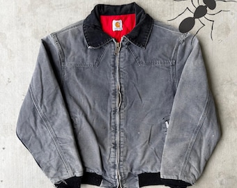 May include: A gray Carhartt jacket with a black collar and cuffs. The jacket has a red interior lining and a zipper closure. The Carhartt logo is visible on the chest. The jacket is a workwear style.