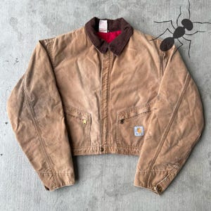 May include: A brown work jacket with a brown corduroy collar and a red interior lining. The jacket has two front pockets with gold zippers and a small square logo patch. The jacket is cropped and has a button closure.