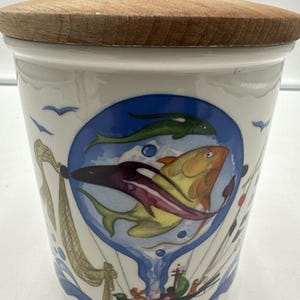 Op de afbeelding: Een witte keramische pot met een houten deksel. De pot heeft een blauw-witte illustratie van vissen, een visnet en een kleine boot. De woorden "AIR FISH" staan onderaan de pot gedrukt.