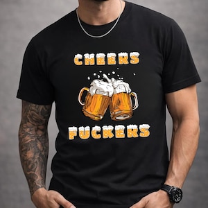 May include: Black t-shirt featuring the text "CHEERS FUCKERS" in yellow, with a beer-foam design. The shirt's central image shows two beer mugs clinking together, with foam spilling over the sides.