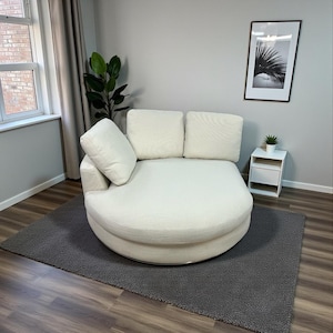 May include: A cream-colored, circular sofa with three back cushions and an angled armrest. The sofa sits on a gray rug, with a small white side table and a framed black and white print in the background. A potted plant is on the side table.