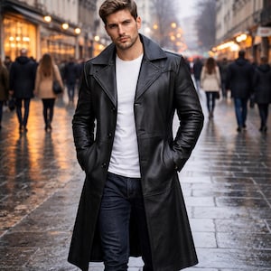 May include: A long black leather coat with a classic collar and button fastening. The coat is worn over a white t-shirt and dark blue jeans. The person is walking on a wet, cobbled street.