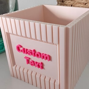 May include: A light pink, square-shaped container with vertical ribbed detailing. The container features the words "Custom Text" in bright pink, raised lettering on the front. The container is empty and sits on a white surface.