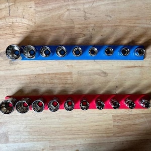 May include: Two socket organizers, one blue and one red, each holding a set of sockets. The sockets are silver and arranged in size order. The organizers are on a wooden surface.