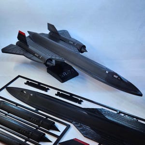 Puede incluir: Maqueta de avión SR-71 negra con detalles rojos en la aleta de cola, exhibida en un soporte con "SR-71". También se ven piezas sin montar, incluyendo alas y fuselaje. El modelo mide aproximadamente 30 cm de largo.