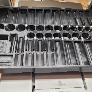 Könnte beinhalten: Eine schwarze Kunststoff-Organizer-Schale mit verschiedenen Fächern und Schlitzen. Die Schale hat rechteckige und kreisförmige Öffnungen unterschiedlicher Größe, die zum Halten und Organisieren kleiner Gegenstände bestimmt sind. Der Text "Ever Of Maker" ist sichtbar.