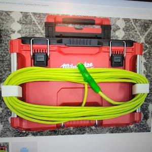 Puede incluir: Una caja de herramientas roja Milwaukee con un cable de extensión verde brillante enrollado alrededor. El cable está sujeto por dos clips blancos. Una caja de herramientas negra y roja más pequeña está encima.