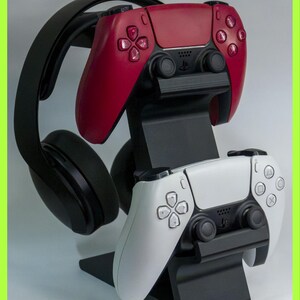May include: A black plastic gaming stand holding a red wireless controller on the top tier, a white wireless controller on the bottom tier, and a pair of black over-ear headphones resting on the side arm of the vertical display mount.