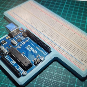 May include: A blue ELEGOO UNO R3 microcontroller board with a breadboard. The board has various electronic components and connectors. The breadboard is white with red and blue lines, and the frame is light blue.