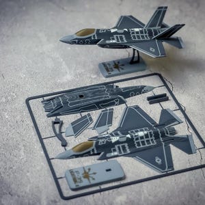 May include: A gray F-35 Lightning II model aircraft kit featuring a fully assembled plane on a display stand and a plastic sprue frame containing unassembled components, including fuselage sections, wings, and a gray base with F-35 text.