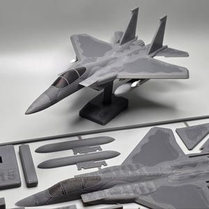 May include: A grey F-15 model aeroplane, assembled and unassembled, on a white surface. The assembled model is mounted on a stand. The unassembled parts are in a grey plastic frame. The text "F-15" is visible on one of the parts.