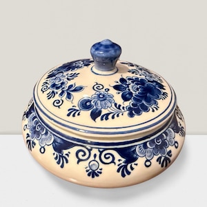 May include: A round ceramic lidded jar featuring a cream-colored base with hand-painted blue floral patterns. The lid includes a central knob handle, and the design displays intricate blue blossoms and swirling leaf motifs across the entire surface.
