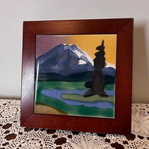 Könnte beinhalten: Eine quadratische Keramikfliese mit einer Landschaftsmalerei, die einen schneebedeckten Berg, einen dunklen Nadelbaum und einen blauen See zeigt, eingefasst in einen dunkel rötlich-braunen Holzrahmen. Der Himmel wechselt von Violett zu Gelb.