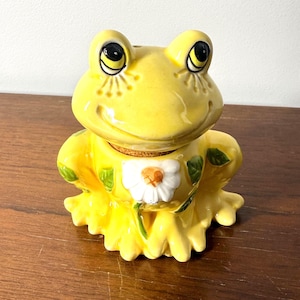 Può includere: Una statuetta di rana in ceramica gialla con finitura lucida, caratterizzata da grandi occhi neri, un'ampia bocca sorridente e una margherita bianca dipinta sul petto. La rana presenta un tappo di sughero sulla parte superiore del corpo.