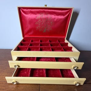 May include: A cream-colored jewelry box with a hinged lid and two pull-out drawers. The interior is lined with red velvet, featuring multiple compartments for organization. The lid interior has a gold floral design, and the drawer fronts feature small, clear, flower-shaped knobs.