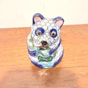 May include: A small cloisonné figurine of a panda bear featuring a white body with blue scale-like patterns, dark blue eye patches, and green bamboo leaves held in its paws. The piece is outlined with thin gold metal wire throughout the design.