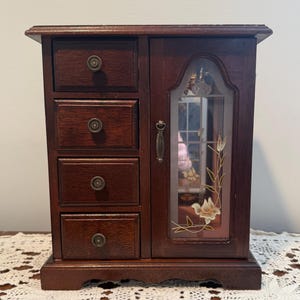 May include: A dark wood jewelry box with four small drawers on the left and a mirrored door on the right. The door is decorated with a floral design. The drawers have round, brass-colored knobs. The jewelry box is sitting on a white lace tablecloth.