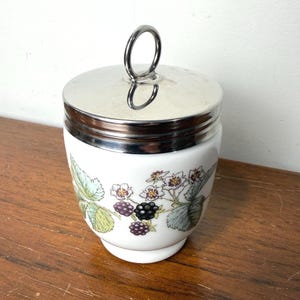 May include: A white ceramic jar with a silver-toned metal lid featuring a circular handle. The body of the jar is decorated with a botanical illustration of green leaves, white flowers, and clusters of dark purple and black blackberries.