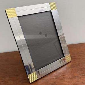 May include: A rectangular silver picture frame with gold-colored accents at each corner. The frame has a black backing and is displayed on a wooden surface. The frame is designed to hold a photograph or artwork.