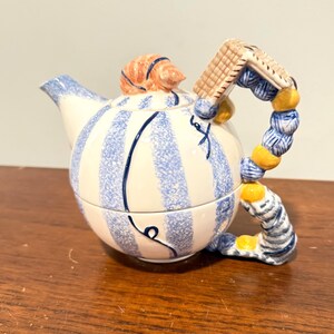 May include: A ceramic teapot featuring a white body with vertical blue speckled stripes and thin dark blue line accents. The lid is topped with a peach-colored seashell, and the handle is shaped like a stack of blue and yellow spheres.