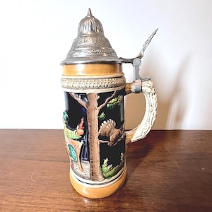 May include: A ceramic beer stein featuring a dark blue body with a relief scene of a tree and a bird. It has a tan base and rim, a decorative metal lid with a thumb lever, and a textured, light-colored handle. The design includes colorful figures.