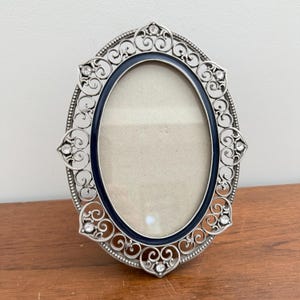 May include: An oval picture frame featuring a silver-toned metal filigree border with intricate scrollwork patterns. The frame includes a dark navy blue inner rim and is accented with small, clear, sparkling crystals set at various intervals.