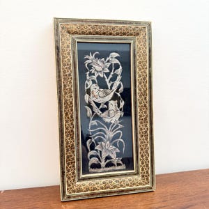 May include: A rectangular wall art piece featuring a black background with an intricate silver-toned inlay of two birds perched among flowers and foliage. The frame is decorated with a dense, repeating geometric star pattern in brown and gold tones.