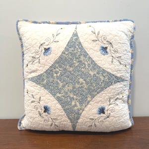 May include: A square quilted throw pillow featuring a central blue and white diamond-shaped paisley pattern surrounded by cream-colored fabric. The cream sections are decorated with embroidered blue flowers and dark green swirling vine motifs.